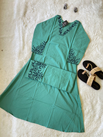 Green dress with black embroidery on a white surface, accompanied by black sandals.