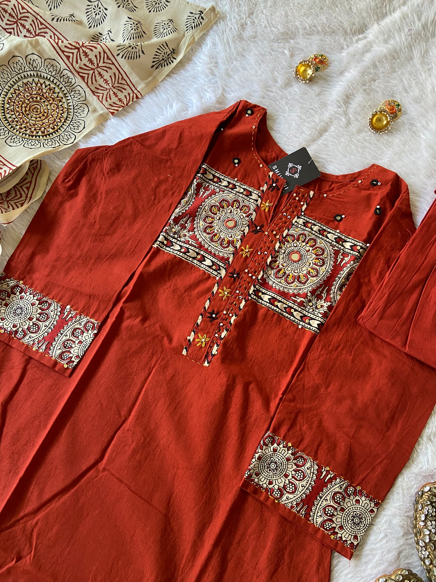 Red embroidered garment on a textured white surface with gold jewelry.