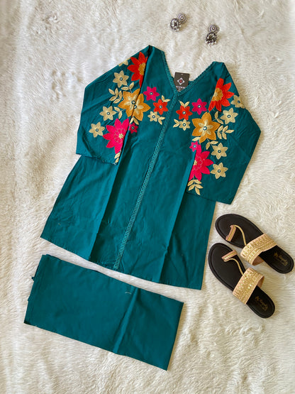 Teal outfit with floral embroidery on a white surface, accompanied by black sandals and jewelry.