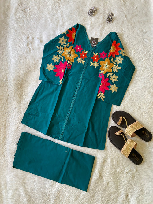 Teal outfit with floral embroidery on a white surface, accompanied by black sandals and jewelry.