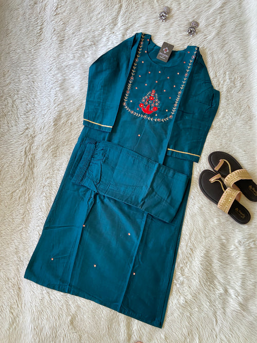 Blue embroidered garment on a textured white surface with sandals in the background