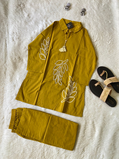 Mustard yellow outfit with leaf patterns on a textured surface, accompanied by sandals and earrings.